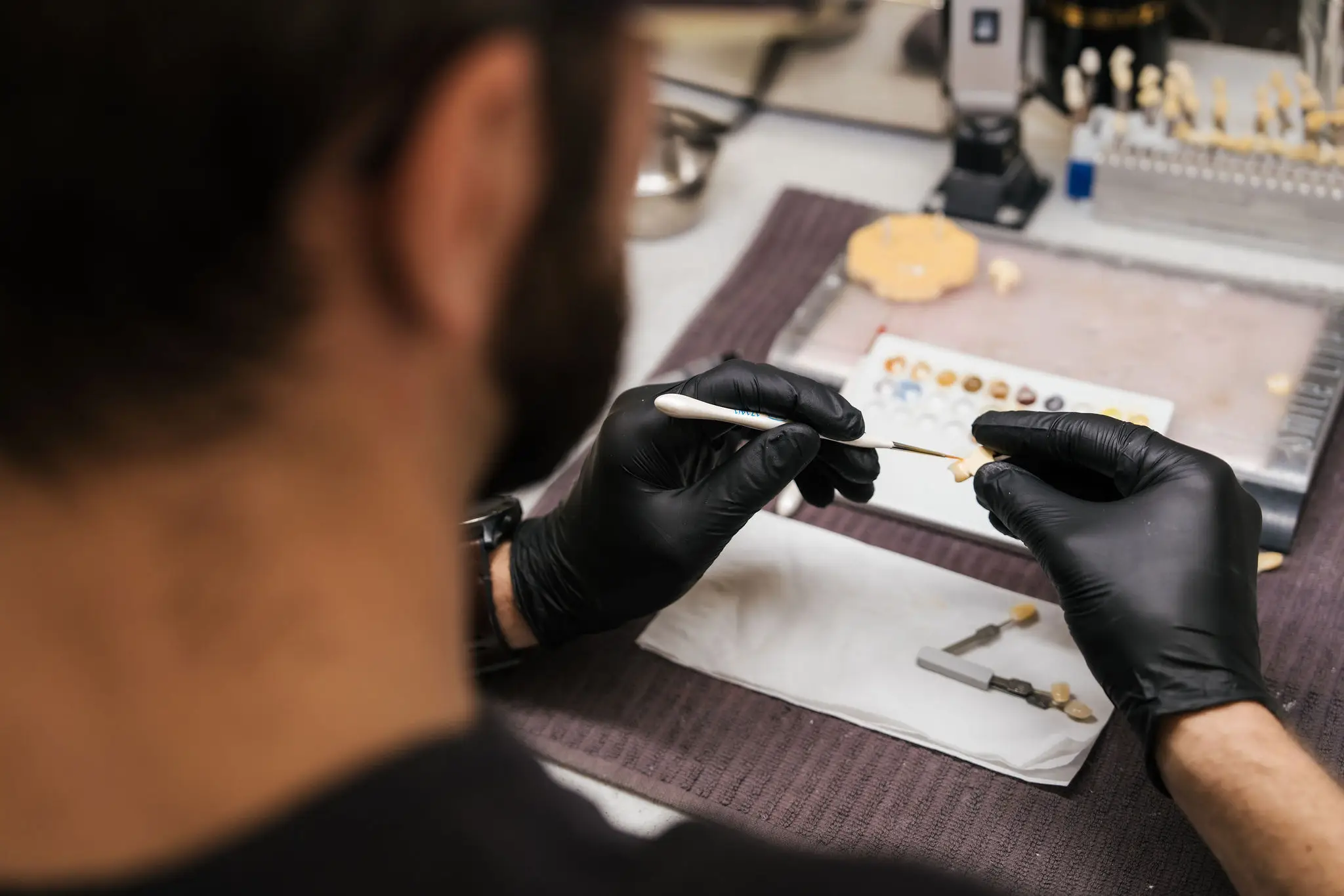 Gloved hands of a dental technician working with tweezers and a fine tool to adjust a small dental component. The workstation is organised with a colour palette, instruments, and materials—capturing a moment of detailed craftsmanship in a clinical laboratory setting.